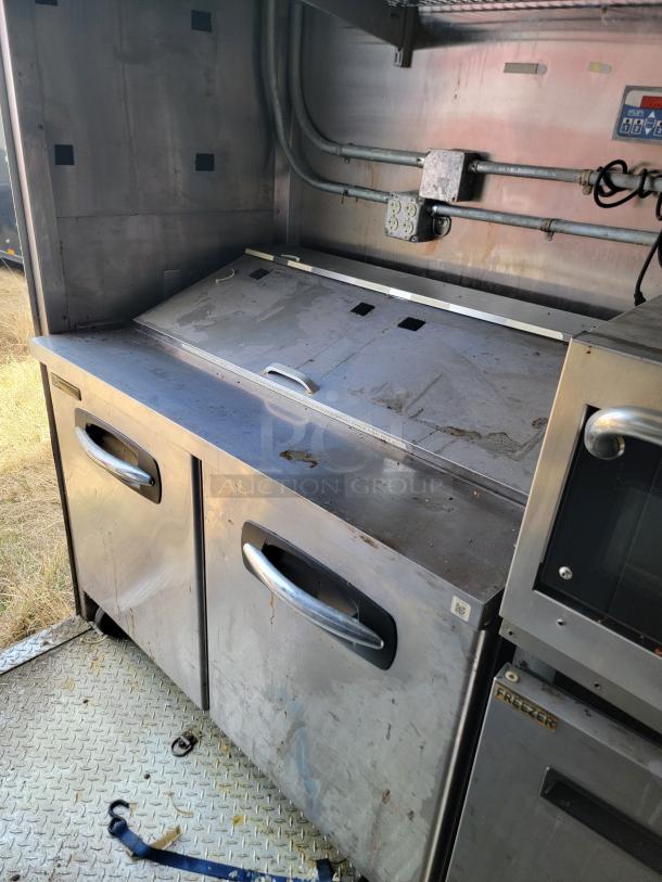 Commercial stainless steel under-counter refrigerator with two doors, pull handles, and visible wear on the surface, ideal for catering.