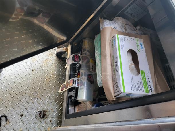 Metal storage compartment with assorted items, including a box of Sysco Reliance vinyl gloves, rolls of stickers, and packaged goods.