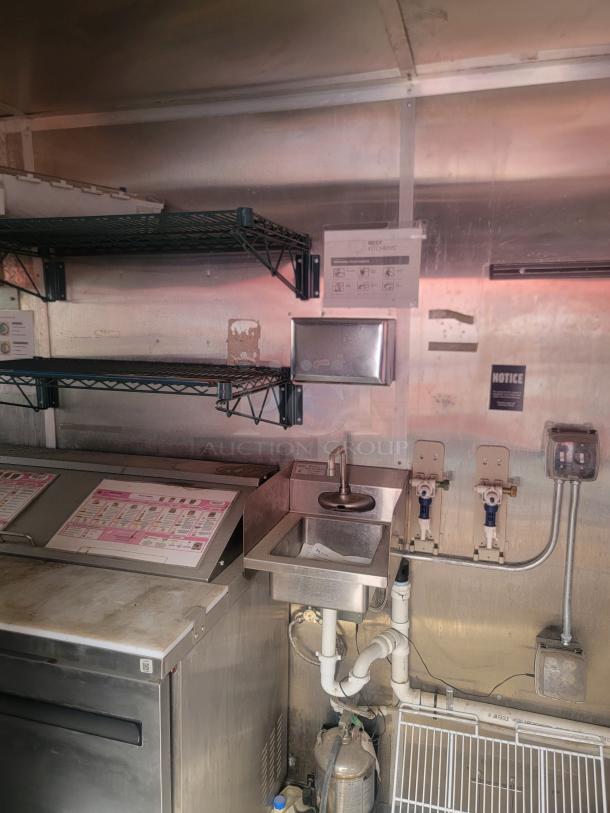 Commercial kitchen setup with stainless steel sink, shelving, and countertop. Visible plumbing and equipment diagrams on the wall.