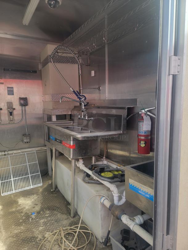 Commercial kitchen setup with stainless steel sink, pre-rinse faucet, storage shelves, and hose reel. Features a fire extinguisher and labeled water line.