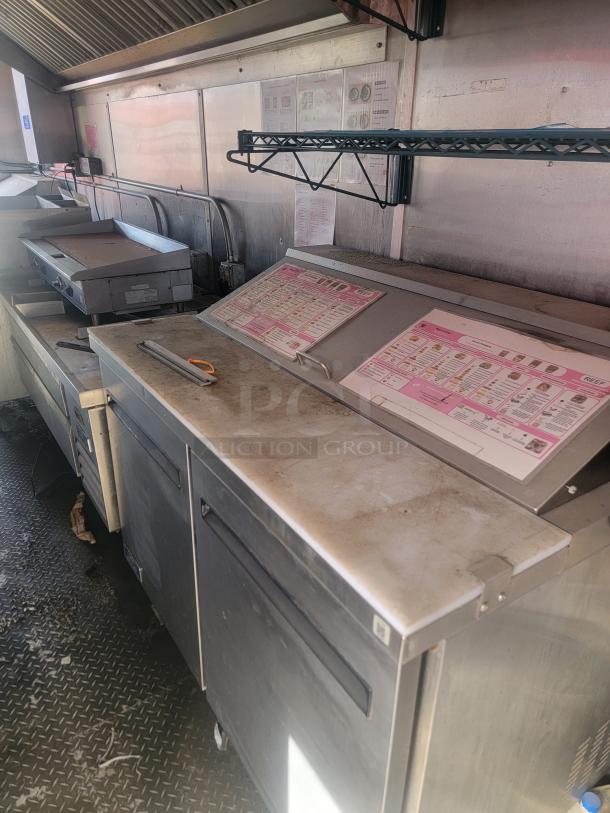Commercial kitchen prep station with stainless steel surfaces, refrigerated storage, menu displays, and sturdy construction. Good condition.