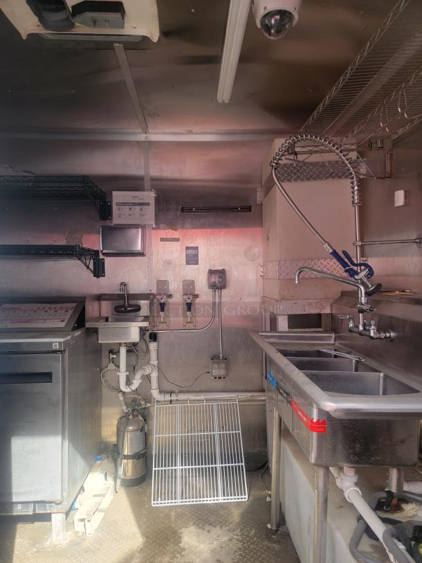 Commercial kitchen interior with stainless steel sink, shelves, and appliances. Visible gas lines and safety notices indicate use.