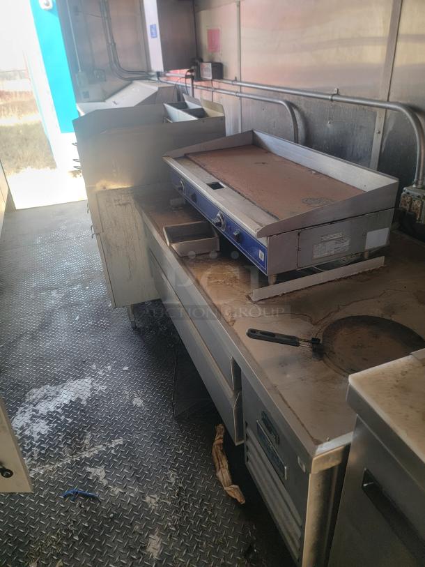 Commercial kitchen grill with two burners, signs of wear. Metal construction with control knobs. Industrial setting, used condition.