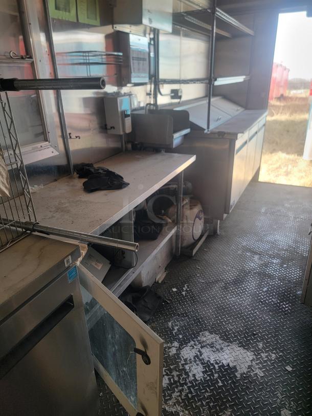 Interior view of a food truck kitchen. Features include stainless steel counters, shelves, a commercial refrigerator, and some open storage. Clean but used condition.