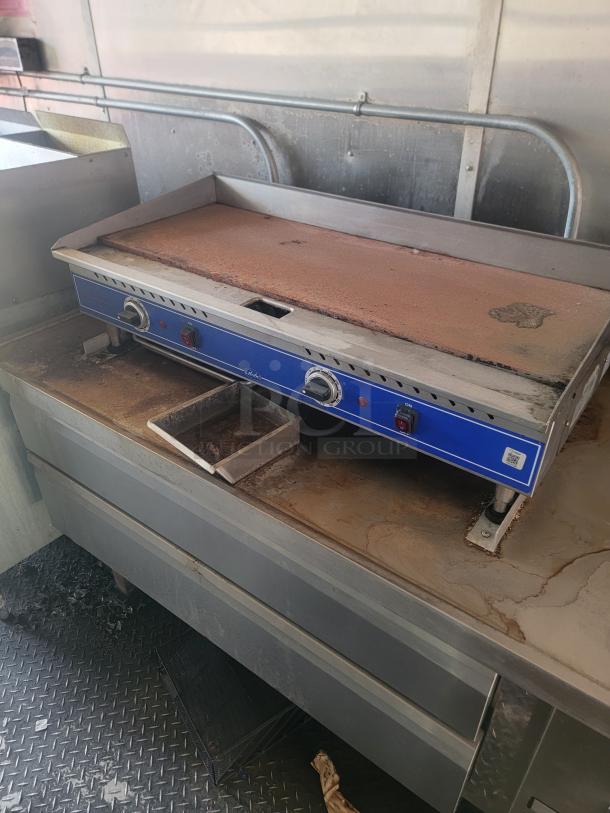 Commercial griddle with blue control panel, dual knobs, on a stainless steel base. Shows signs of use, includes grease tray.