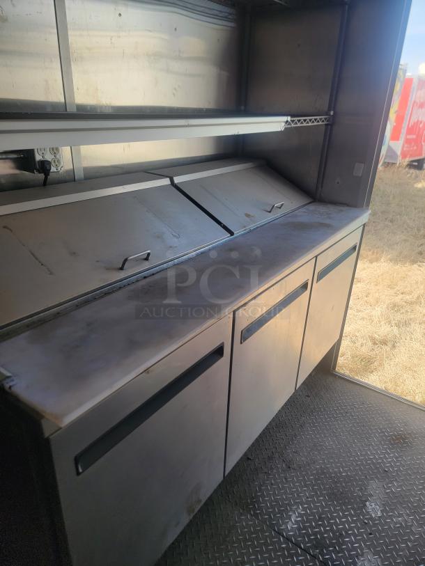 Stainless steel commercial prep table with storage units and drop-down lids. Shows signs of use, no visible markings.