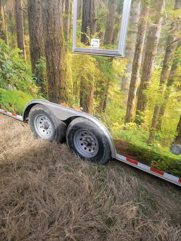 Enclosed trailer with forest-themed exterior wrap, dual axles, silver rims, and reflective red-white trim. In grassy area.