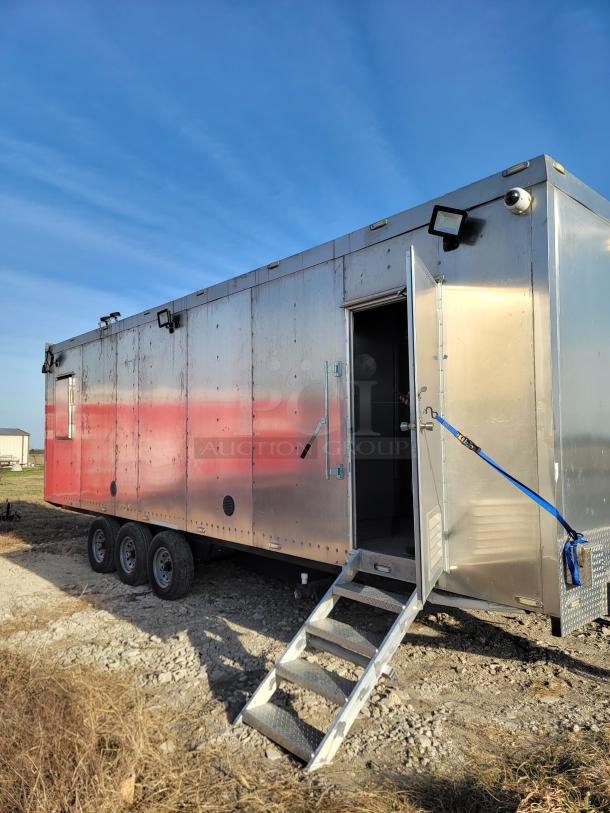 Silver mobile trailer with red accents, featuring triple axles, a side door with metal steps, and mounted security cameras.