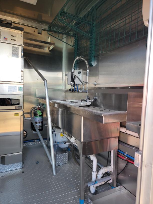 Commercial kitchen interior in a food truck, featuring a stainless steel sink with a high-arch faucet, shelving, and industrial equipment.
