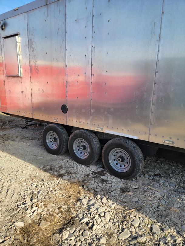 Metallic travel trailer with triple-axle wheels, weathered exterior, and side vent; parked on rocky ground.