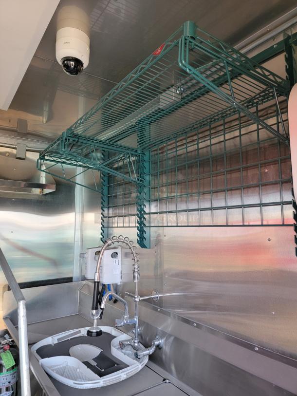 Commercial kitchen sink unit with metal shelving, wall-mounted sprayer, and visible security camera, in excellent condition.