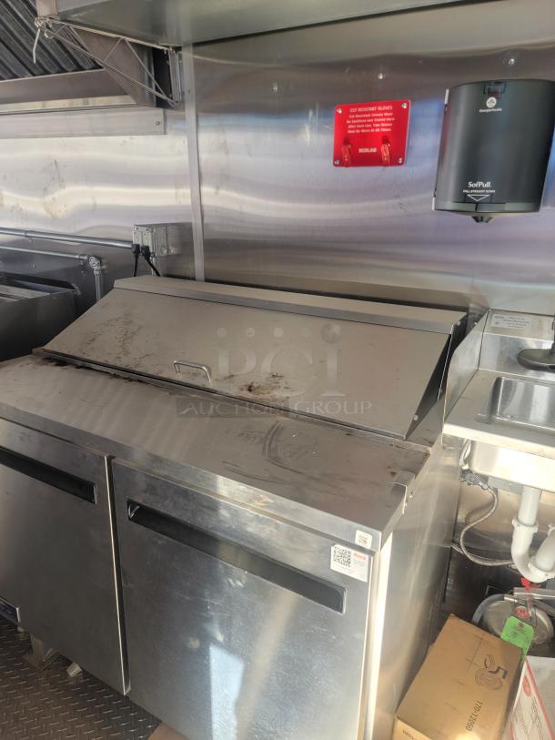 Stainless steel commercial refrigerator with two doors, showing signs of wear. Visible QR code and "Ecolab" glove sign above.