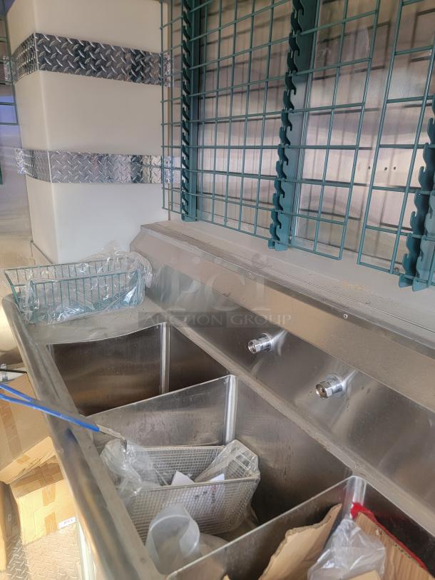 Commercial stainless steel triple sink with two faucets, metal grid back, and storage shelves. Appears in good condition.