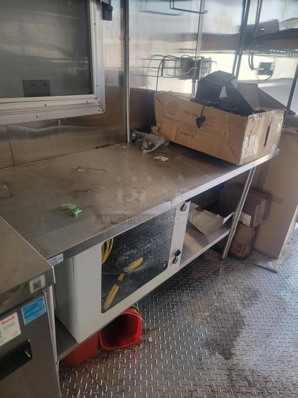 Stainless steel worktable with storage shelves, minor wear. Features side cabinet with transparent door, visible wires, boxed items.