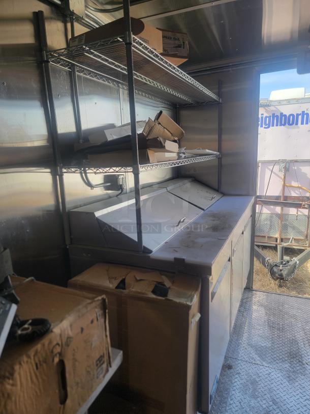 Commercial kitchen shelving and refrigerated prep table. Stainless steel, shows wear. Cardboard boxes stored on shelves.