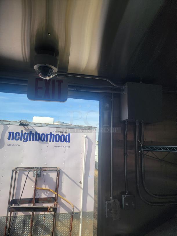 Ceiling-mounted dome camera near an "EXIT" sign inside a trailer with visible wiring and exterior view of a "neighborhood kitchens" trailer.