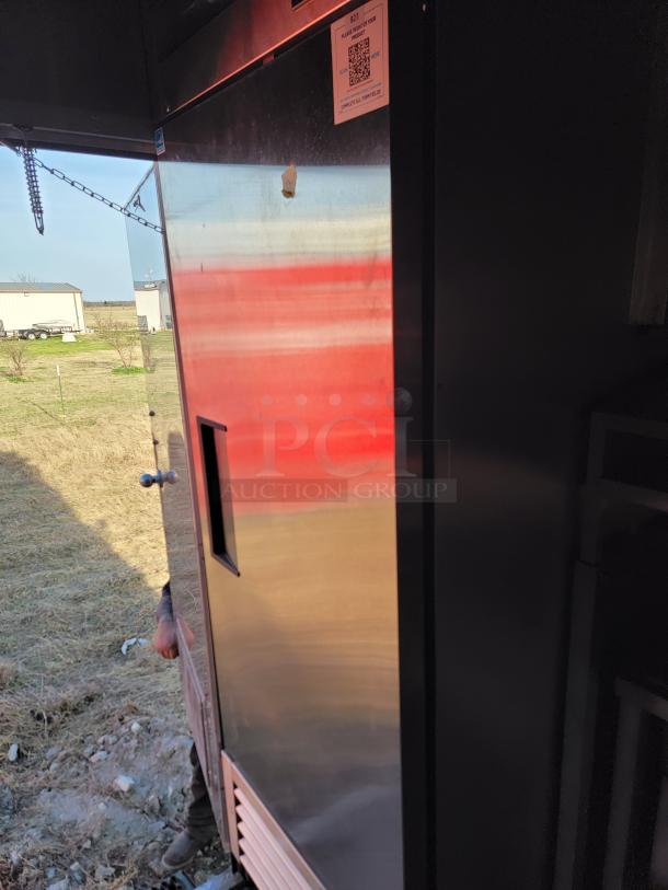 Commercial stainless steel refrigerator, used, with external handle and visible wear. QR code label on top corner.