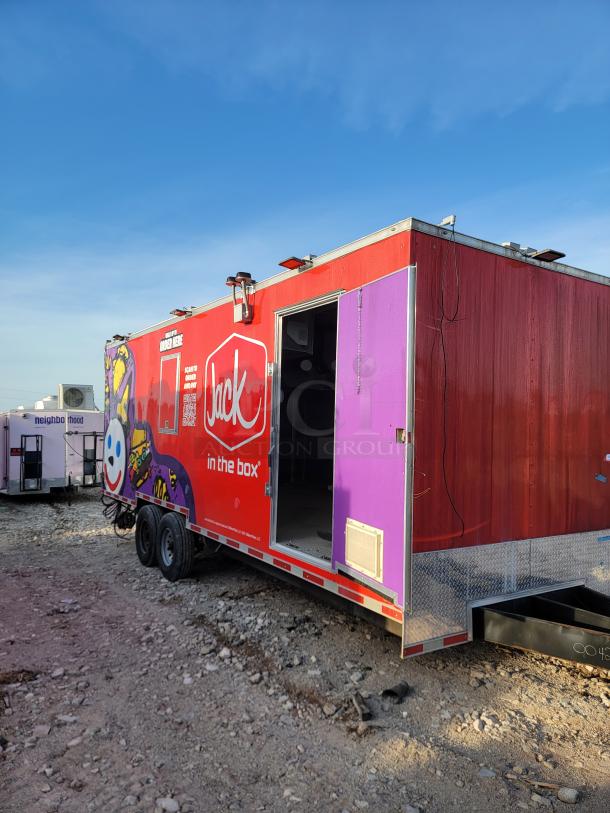 Red and purple "Jack in the Box" food trailer with logo and graphics, open door, visible wear, parked on gravel.