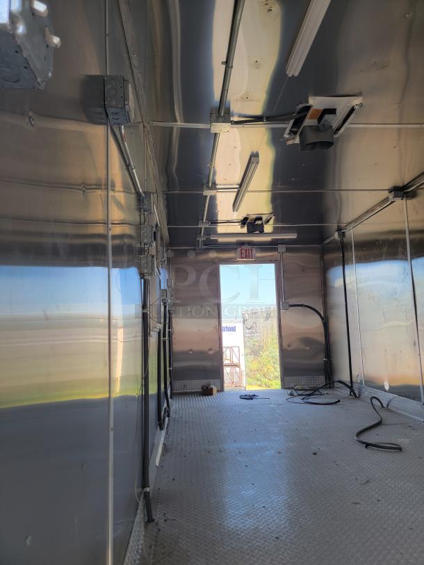Interior view of a metallic trailer with a textured floor. Visible wiring and fluorescent lights installed. Exit sign above open rear door.
