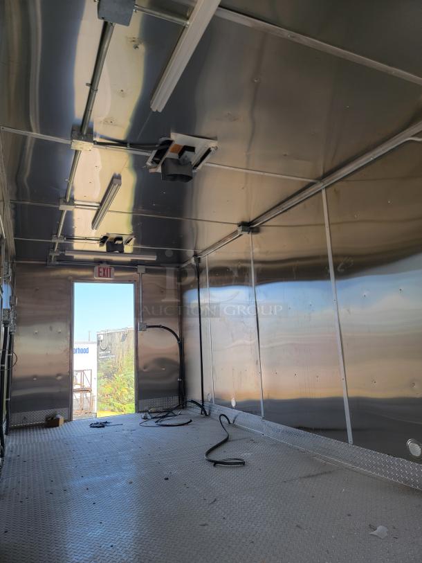 Empty metal trailer interior with diamond plate flooring, wiring, and fluorescent lighting. Exit marked at end. Clean condition.