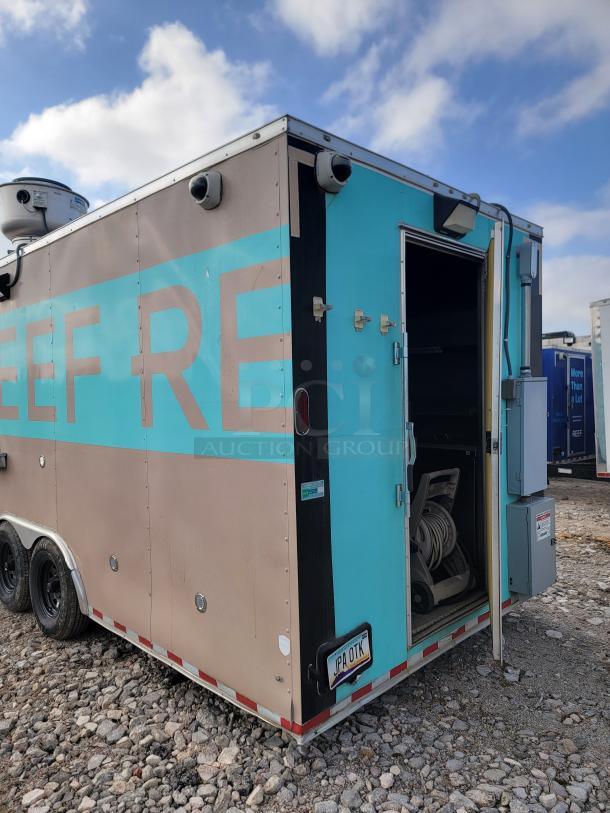 Blue and beige mobile service trailer with security cameras, open side door, dual axles, and visible equipment inside. License plate marked "JPA 01K."