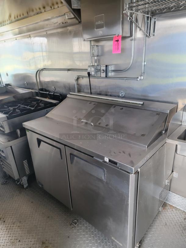 Stainless steel commercial refrigerator with two doors, shelf above, visible wear, pink maintenance tag attached. Ideal for kitchen use.