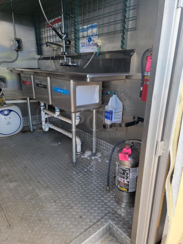 Commercial stainless steel triple sink with faucet, labeled compartments, safety equipment, and cleaning supplies in used condition.