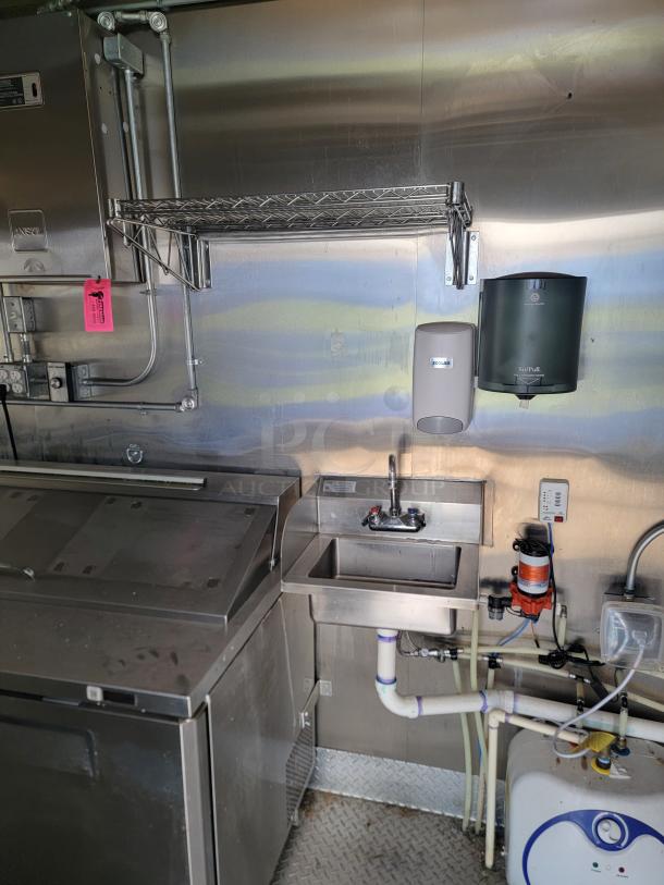 Commercial stainless steel kitchen setup with sink, wall rack, dispensers, and plumbing. Contains utility stickers and labels.