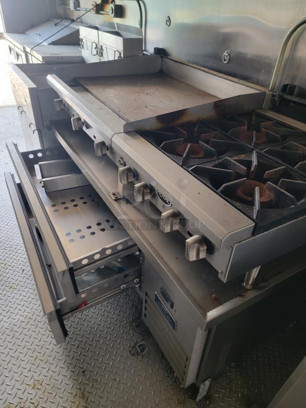 Commercial stove and griddle unit with six-burner cooktop and storage drawers. Stainless steel finish, signs of use visible.