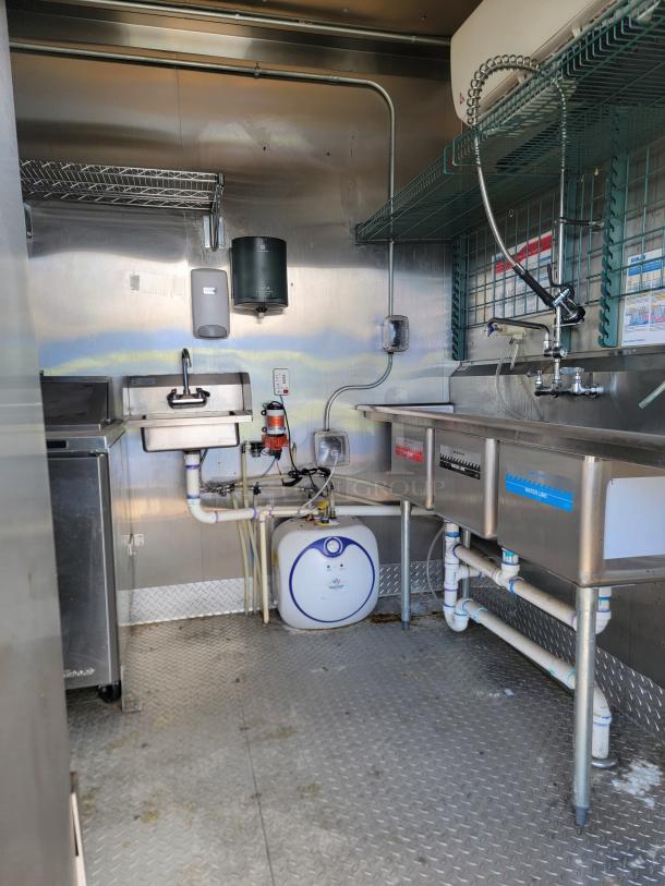 Commercial kitchen setup with stainless steel sinks, faucet, shelves, plumbing, water heater, and wall-mounted dispensers.