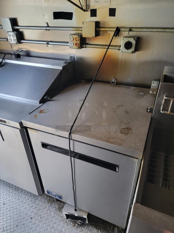 Commercial stainless steel refrigerated prep table, top surface shows signs of wear and stains, visible power cord and outlet.
