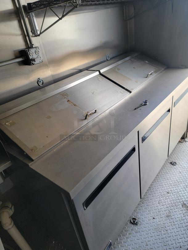 Stainless steel commercial refrigerator with dual top lids, visible handles, minor wear, integrated storage; ideal for kitchen use.