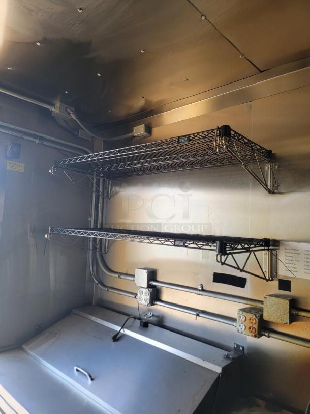Commercial stainless steel shelving unit mounted on a food truck wall, featuring dual wire racks and visible electrical outlets.