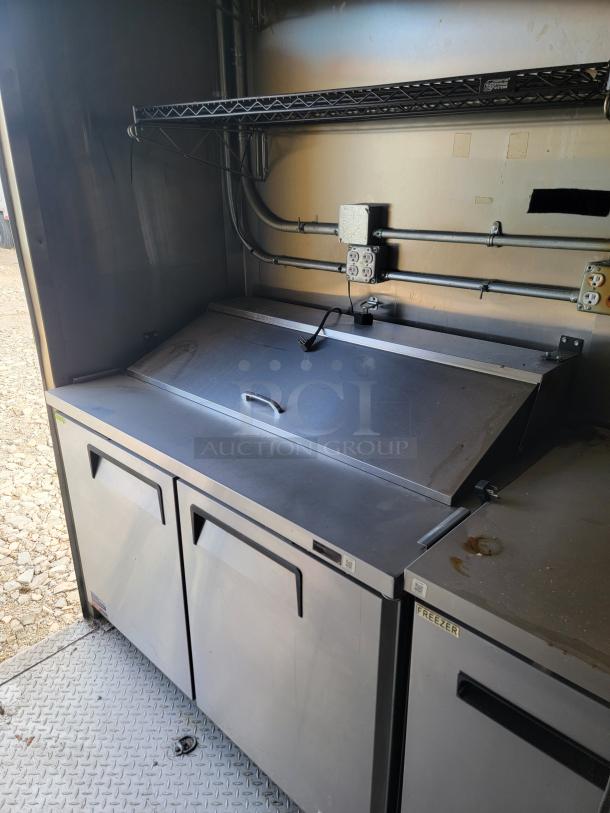 Commercial stainless steel refrigerated prep table with dual storage doors, a hinged top, and visible power outlets. Good condition.