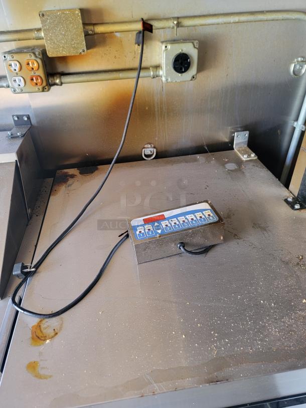 Industrial control panel with attached cables on a metal surface, showing signs of wear and rust. Includes labeled buttons and dials.