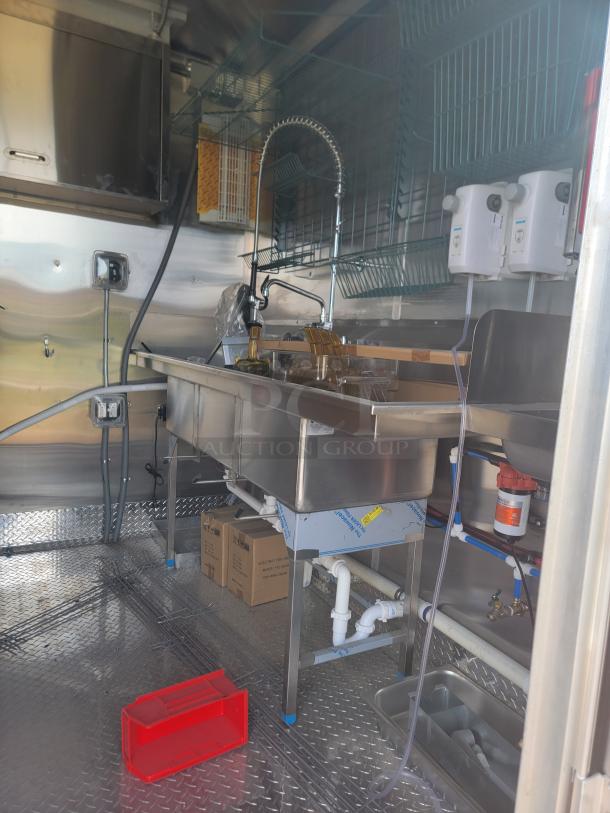 Stainless steel commercial kitchen setup in a mobile unit, featuring a deep sink with a high-arc faucet, wire racks, and plumbing fixtures, all in pristine condition.