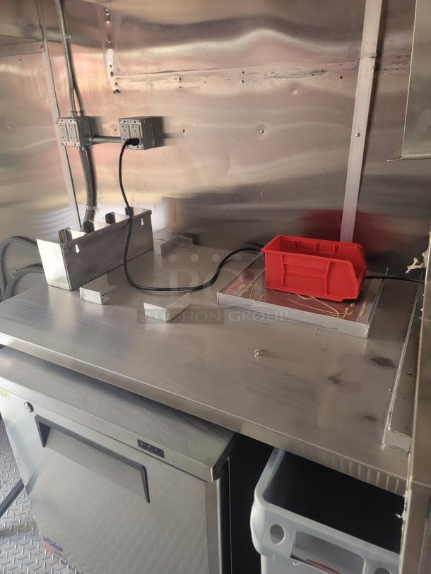 Stainless steel workbench with two power outlets, a small under-counter refrigerator, and a red storage bin.