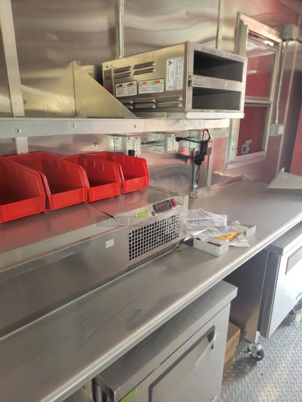 Commercial stainless steel food prep station with warming unit and storage, featuring red bins, shelving, and paperwork.