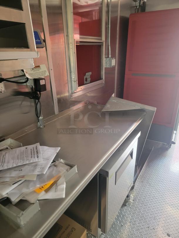 Interior of a food truck with stainless steel counter, undercounter fridge, and wired shelving; papers and manuals visible.