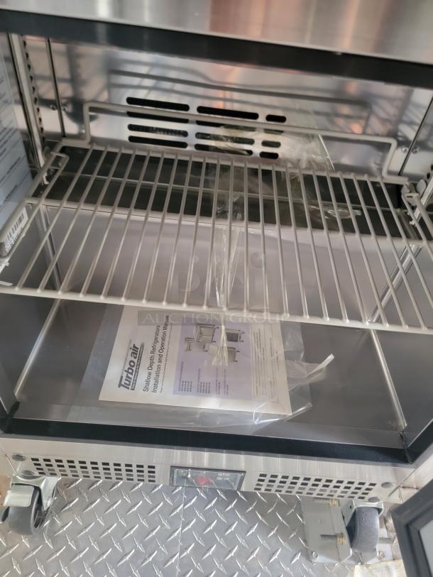 Turbo Air refrigerator interior, stainless steel, wire shelf, manual included, on wheels. Features control panel and ventilation.