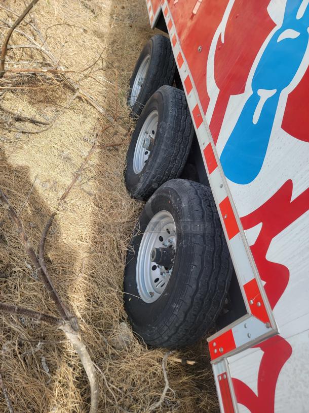 Red and white trailer side with three black tires on dry grass; visible blue graphic on panel.