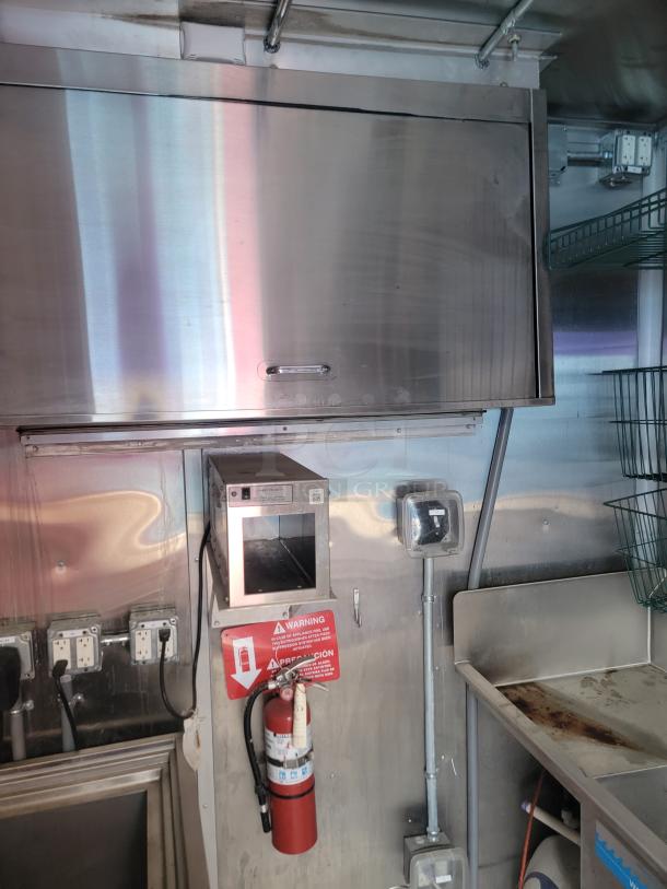 Stainless steel commercial kitchen setup with wall-mounted exhaust hood, electrical outlets, fire extinguisher, and storage shelf.