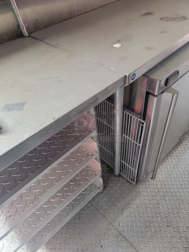Stainless steel commercial kitchen countertop with shelves and NSF-certified label, showing signs of wear.