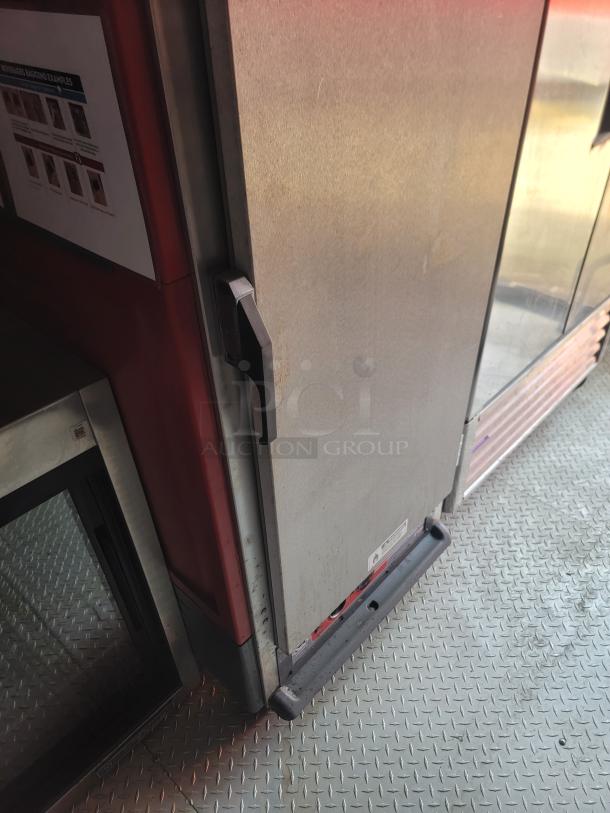 Commercial metal refrigerator with black handle, slight wear, on diamond plate flooring. A visible label indicates model details.