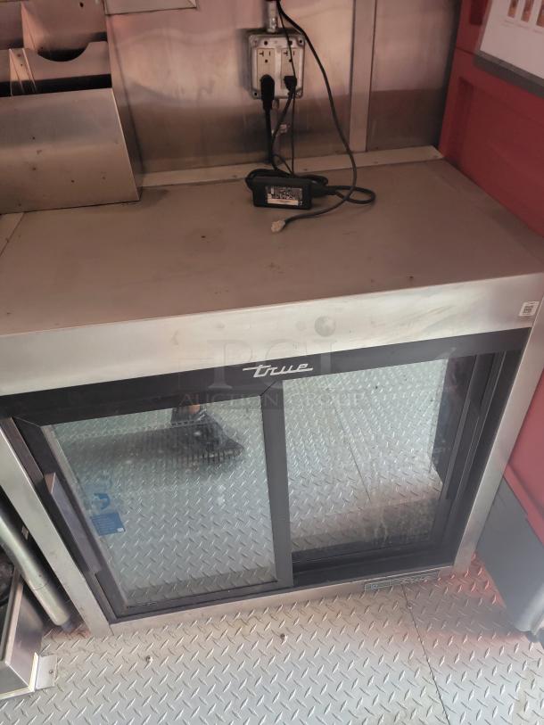 True commercial beverage cooler with glass doors, stainless steel top, black trim, visible power cords, and "True" logo.