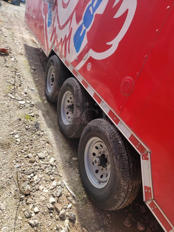 Red trailer with triple axles and steel wheels on a gravel path. Features a large, colorful graphic on the side. Good condition.