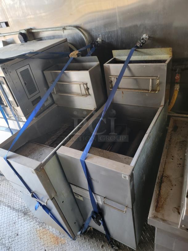 Commercial deep fryer units with metal baskets, secured with blue straps, showing wear and signs of use, in stainless steel finish.