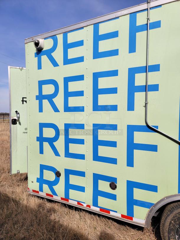 Green mobile unit with "REEF" in blue letters, exterior surveillance camera, and ventilation ports. In grassy field.