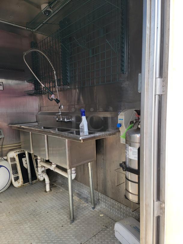 Stainless steel commercial sink with sprayer and three basins, mounted in a metal-lined area. Includes a wall rack and Ecolab soap dispenser.