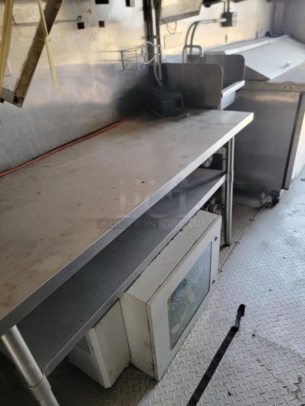 Commercial kitchen workspace with metal countertop, lower shelf, and equipment beneath. Visible wear, stainless steel.